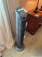 Front view of the gray Seville Classics tower fan standing on a carpet next to a wooden nightstand and curtains.