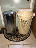 Two items side by side on tiled floor: shiny metal garbage can with pedal lid and beige plastic laundry hamper with ventilated design and lid, placed on a half-circle patterned mat.