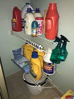 Photo showing two shelves with various bottles including Resolve, Springtime fabric softener, Arm & Hammer Essentials detergent, ZEP carpet cleaner, SoftSoap, Old Dutch, with spray bottles hanging and bucket below.