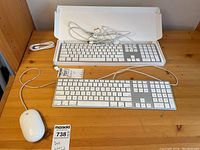 Apple MB110LL/B wired keyboards and a white Apple wired mouse arranged on a wooden surface, keyboards show wired connection cables, one keyboard is in original packaging box.