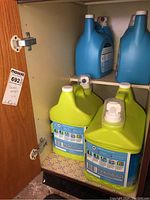 Four large bottles inside cabinet: two green Springtime detergent bottles in front lower shelf, two blue Downy fabric softener bottles on upper shelf.