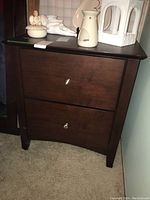 Photo of one dark wood nightstand showing two drawers with silver round knobs and items placed on top.
