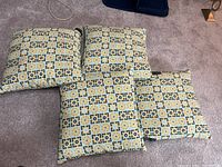 Four outdoor cushions with a patterned design in shades of yellow, blue, black, and white laid on carpet.