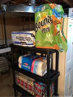 Photo showing three shelves of paper goods in basement setting: Kirkland Bath Tissue, Bounty Plus Paper Towels, Cashmere Deluxe Toilet Paper, and Scotties facial tissue boxes with floral/beige design.
