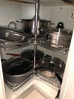 Corner cabinet with multiple pots and pans stacked on metal wire shelves, mostly stainless steel with lids.
