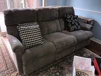 Front and side view of a three-seat gray-beige upholstered powered reclining sofa with plush backrest and armrests, each end reclines, middle seat fixed. Sofa includes two black and white circular pattern cushions and one black and white leaf pattern cushion.