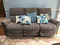 Full view of gray upholstered powered reclining sofa with three seating sections, middle does not recline. Five decorative throw cushions on sofa, three floral and two blue quilted.