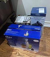 Overview of all three original boxed items: Oster hand blender in blue box, Trudeau stainless steel pump carafe in blue box, Black & Decker cordless kettle in black and white box