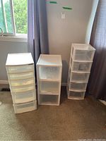 Wide shot of three plastic storage drawer units positioned against a wall near a window and curtain; varying sizes with drawers visible.