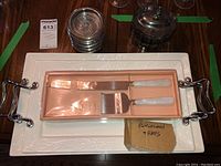 Two white porcelain serving trays stacked, each with metal handles and embossed leaf detailing. Cake serving set in pink box on top.