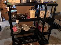 Two black plastic utility shelving units placed side by side with various household items on shelves (items not included in lot).