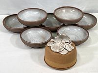 Seven hand-formed stoneware bowls with glazed interiors in light gray with concentric pattern, and reddish-brown unglazed exteriors, arranged stacked and beside ocean-themed covered dish.