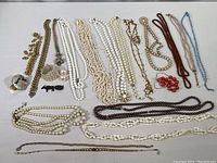 Full view of the jewelry collection spread out on a white surface, showing mostly necklaces, a compact item, earrings, pin, and bracelet.