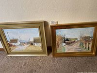 Two framed landscape paintings leaning against wall showing coastal house scenes.