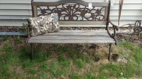 Full view of outdoor bench showing wood slatted seat, decorative cast iron back with floral pattern, and armrests. One pillow on bench for size reference.