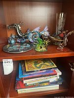 Shelf with various dragon figurines including art glass and metal, collector's plate and stack of dragon-themed books beneath.