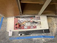 Overview photo of all components: miter gauge, black metal fence with handle, and a plastic bag with accessories on wood shop table.