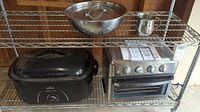 Photo of entire lot on metal wire shelf: Rival Roaster Oven on bottom left shelf, Cuisinart Toaster Oven Grill on bottom right shelf, metal bowl with lid, metal pan lid, and metal mug on top shelf.