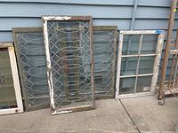 Five salvaged wooden windows with latticed and paned glass propped against a blue wall on concrete, showing weathered wood and chipping paint.
