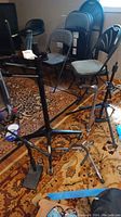 Two black metal Gator Frameworks music stands on patterned rug, with tripod bases and clamps, surrounded by chairs and room items.