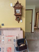 Vintage wall clock visible on wall above stack of moving boxes and a black chair holding the wooden box with vase.