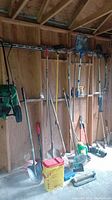 Photo showing an assortment of garden tools including rakes, shovels, a container, and other gardening supplies hung or leaning against a wooden wall in a shed.