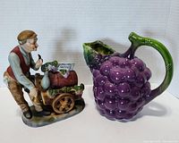 Photo showing bisque porcelain Veno cart vendor figurine next to grape bunch pitcher on white background.