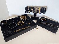 Photo showing all black and gold-tone bracelets and bangles arranged on black trays and a bracelet stand.