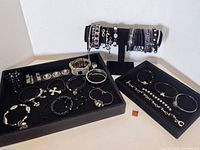 Full view of all costume bracelets and bangles displayed on two black velvet trays and one stand, showcasing the silver and black color tones and varying bracelet styles.