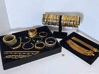 All bracelets arranged on black display stands and flat surfaces, showing variety of gold tone costume bracelets and bangles, including thick cuff, hoops, chain styles, and multi-strand bracelets.