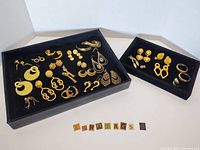Photo shows two black trays holding a variety of gold tone costume pierced earrings displayed in neat rows, showing different styles including hoops, studs, and drop earrings.