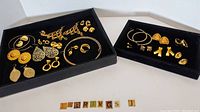Lot of various gold tone pierced earrings displayed in black trays, showcasing multiple pairs of earrings including large hoops and geometric shapes.
