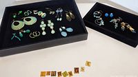 Lot of pierced costume earrings shown in two trays, displaying varieties of green, teal, and blue tones, multiple styles including studs, hoops, and dangles.