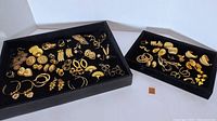 Wide view of two black trays with multiple pairs of gold tone pierced costume earrings in various styles and sizes.