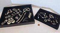 Overview of mostly silver tone pierced earrings in two black trays showing multiple pairs with various designs including hoops, chains, discs, and shaped danglers.