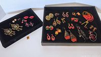 Two trays with multiple pairs of costume earrings in reds and purples, showing variety of styles and sizes.