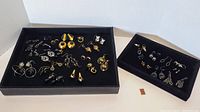 Overview photo showing two black trays with many pairs of black and gold-toned pierced costume earrings of varying styles including studs, dangles, hoops, and chandeliers.