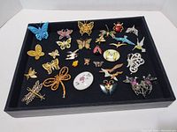 Front angled view of all 28 pins and brooches laid out on black display tray showing insect and butterfly themed costume jewelry