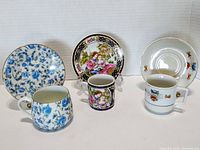 View of all three tea cup and saucer sets showing floral blue design, romantic couple scene with black and gold decoration, and white small flower design.