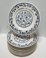 Stack of 10 white ceramic dinner plates with blue floral Sturbridge pattern on front, displayed on clear plate stand.
