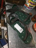 Pair of green aerating shoes with large metal spikes and black adjustable straps for attaching to footwear placed on metal surface with vintage oil cans in background.