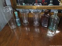 Six antique glass bottles and beakers arranged on a wooden surface reflecting in a mirror. Varied sizes and colors including clear, blue-green, and brown glass.
