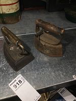 Two antique irons placed side by side, showing the wood-handled iron with a hinged removable cover and the other iron with a fixed metal handle, both exhibiting rust and aged wear.