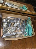 Full view of tray filled with assorted older metal cutlery including spoons, knives, and other utensils alongside a greenish-blue cloth pouch.