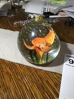 Side view of vintage glass paperweight showing embedded orange flower design with green leaves and air bubbles inside clear glass.