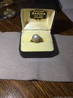 Sterling silver ring with large white pearl-like stone inside a black ring box with yellow interior labeled 'Bijouterie Sabra, 1453 Peel, Montreal'.