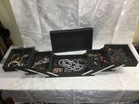 Full view of the multi-level black folding jewelry case showing various trays filled with vintage jewelry including necklaces, earrings, bracelets, pins, and rings.