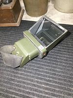 Side and angled view of a vintage military tank periscope showing large angled glass viewer and olive green metal body with rubber eyepiece.