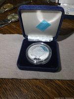 Silver coin in protective plastic capsule displayed inside original blue velvet box with white satin lining, view including sticker with numbers on inside lid