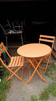 Photo of orange folding metal bistro table and two matching chairs set up outside.